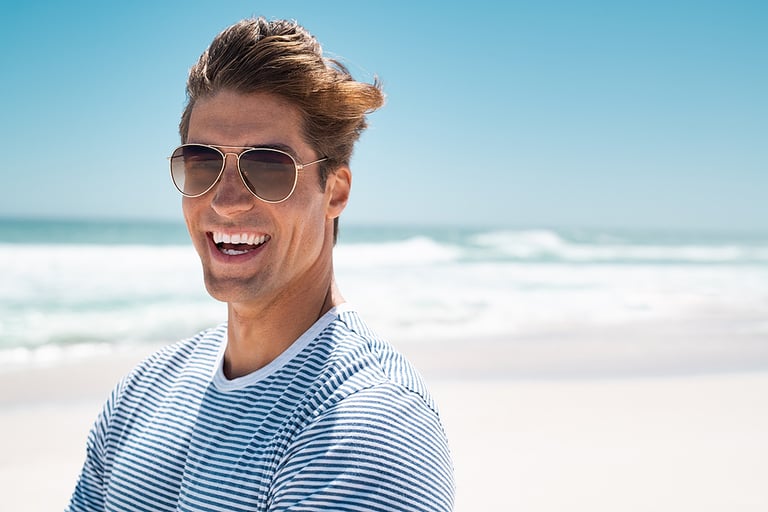 Portrait of handsome young man wearing sunglasses and looking at camera. Portrait of joyful man enjoying summer holiday at beach. Young casual man in shades with big grin and copy space.
