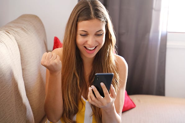Happy overjoyed young woman holding mobile phone celebrating bid win game app victory sit on sofa at home, excited euphoric student girl winner rejoicing success with cell phone