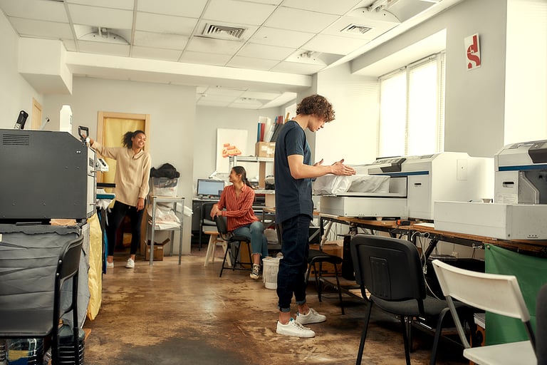 Full-length shot of young cheerful millennials using heat press transfer, silk screen printing machine for printing t-shirts at workplace. Printing on clothing and textiles concept. Horizontal shot