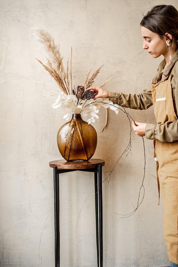 Woman decorating home with a composition of dried flowers and herbs in a glass vase on a beige wall background