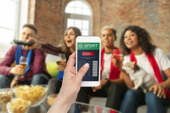 Device screen with mobile app for betting and score. Device with match results on screen, excited fans on background during match. Gambling, betting, sport, finance, modern technologies concept.