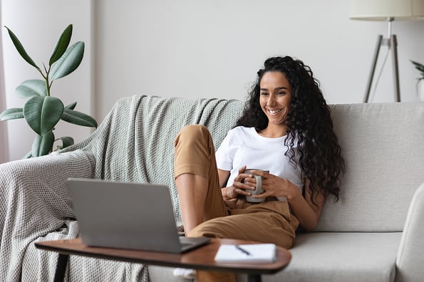 Relaxed pretty young woman watching movie on laptop, drinking coffee, sitting on sofa at home, copy space. Happy curly lady enjoying her weekend alone, watching comedy online, laughing