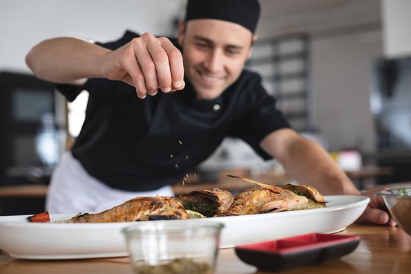 Male caucasian chef adding salt over roasted fish dish in food plate at restaurant kitchen. food preparation restaurant culinary gourmet