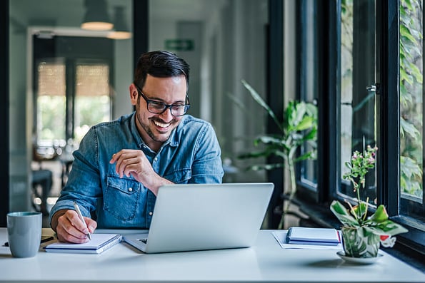Happy male entrepreneur writing ideas on note pad. Businessman working on laptop at table. He is doing freelance work at home office.
