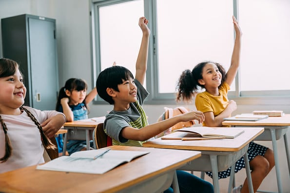 Elementary age Asian student boy raised hands up in class. Diverse group of pre-school pupils in elementary age in education building school. Volunteering and participating classroom concept.
