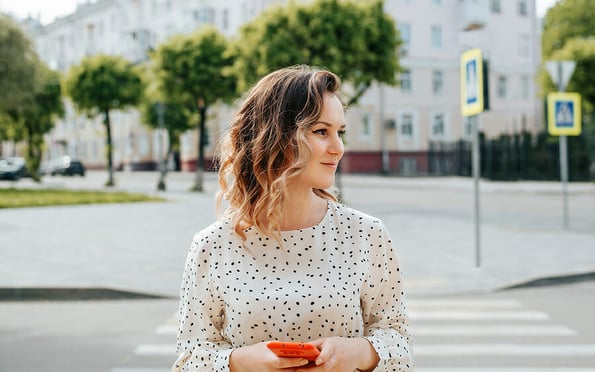 Stylish smiling young woman crossing crosswalk and looking away. Portrait of smiling pretty woman with curly hair holding mobile phone and walking along road in city.