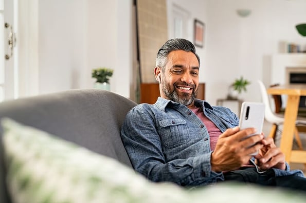 Happy mid adult man using smartphone device while sitting on sofa at home. Smiling mature indian man lying on couch reading messages on mobile phone while listening to music with wireless earbuds.