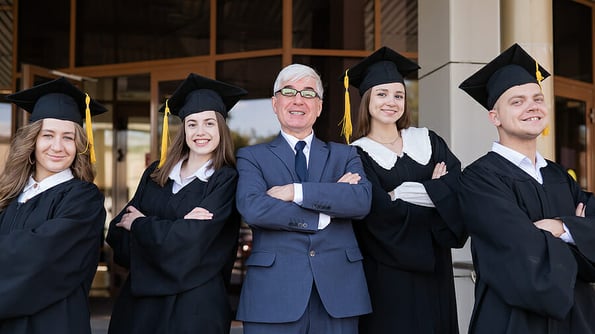 A professor and his students in graduation gowns stand with their arms crossed over their chests.