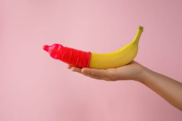 Woman holding banana in condom on pink background, closeup. Safe sex concept