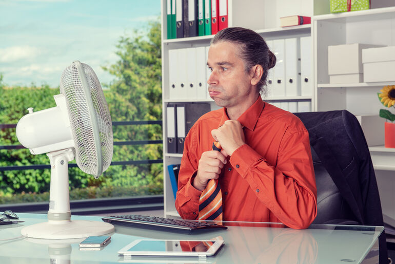 Office worker sitting at a desk in front of a fan, looking uncomfortable and overheated despite indoor cooling, illustrating a muggy office environment.