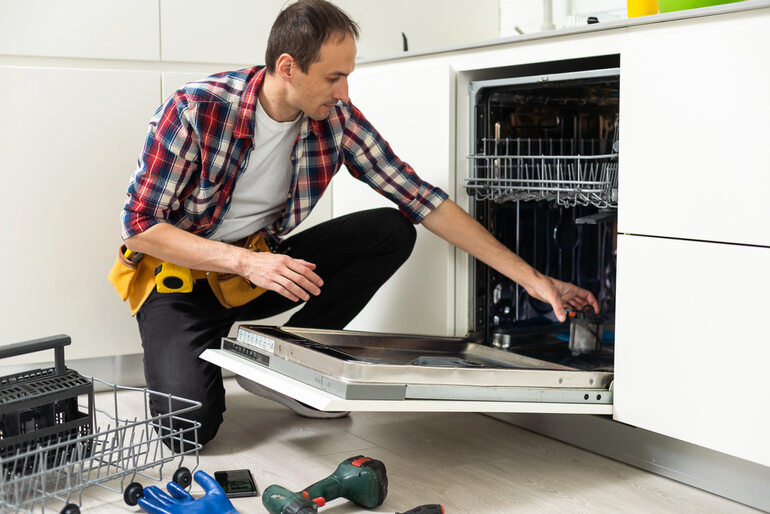 A man sitting on the floor carefully cleaning the coils of a refrigerator, focusing on proper maintenance to improve appliance efficiency and performance.