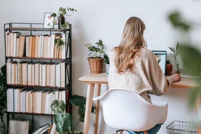 Adult woman working on a laptop at a home office desk surrounded by a corner bookshelf and plants
