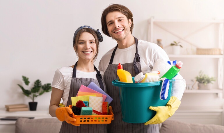 Couple working together on household responsibilities, with one carrying cleaning supplies and the other holding a basket of household items.