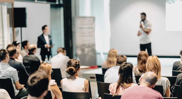 Audience seated at a business conference listening to a speaker presenting in front of a screen.