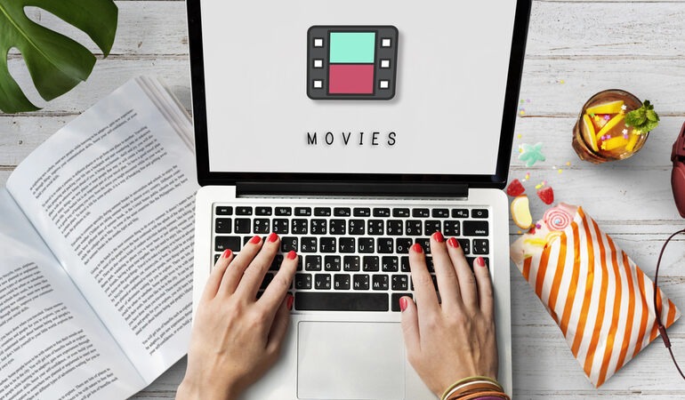 Person browsing movie entertainment on a laptop beside an open book, popcorn, and a drink.