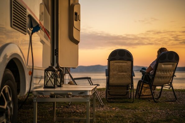 Scenic RV Campsite Pitch. Camper Van In the Recreational Vehicles Park. Woman Relaxing on a Chair and Watching Sunset Over the Sea.