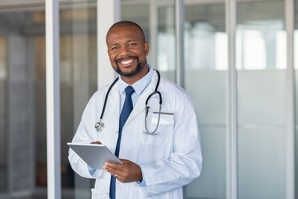 Portrait of confident mature black doctor consulting digital tablet and looking at camera. Smiling african american doctor with stethoscope using tablet at medical clinic. Happy healthcare worker.