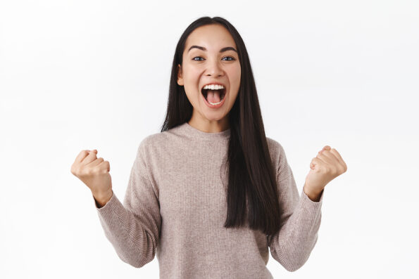 Excited and happy cheerful asian woman clench fists in powerful triumphing motion, fist pump and shouting yes from amazement and joy, smiling pleased, achieve success, receive prize, winning lottery