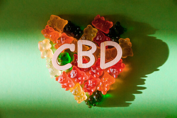 Photo of Wooden letters CBD on a green background and bears gelatin in the form of heart. View from above. The shadow of the sun falls on the background