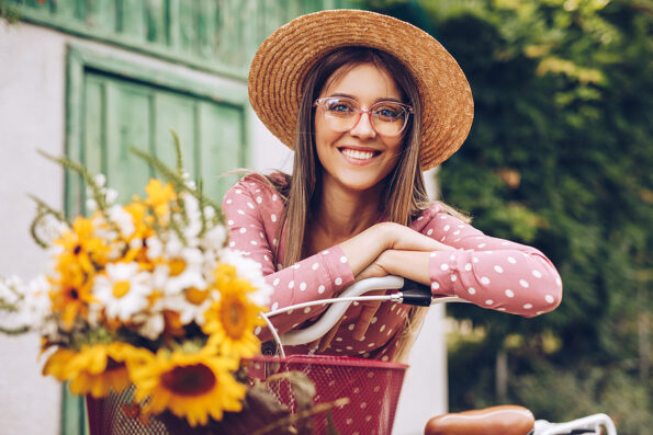 Optimistic young woman in stylish hat and glasses smiling for camera and leaning on bicycle with flowers, while resting in park on sunny day in summertime