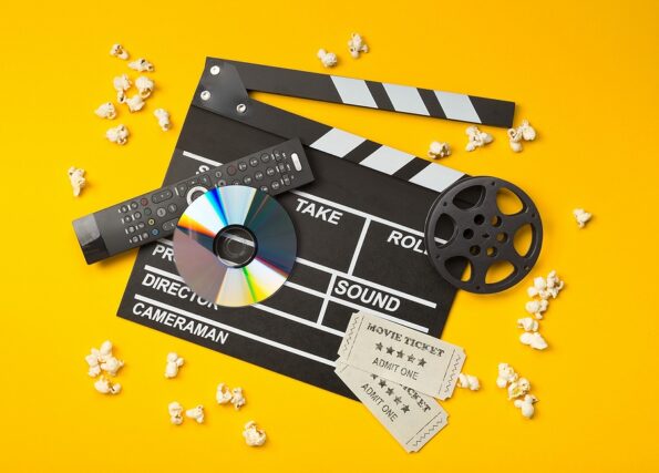 Single, black, open movie clapper or clapper-board with dvd movie disc, film reel, popcorn, remote control and movie theatre tickets flat lay top view from above on yellow or orange background - digital movie, home cinema or movie night concept