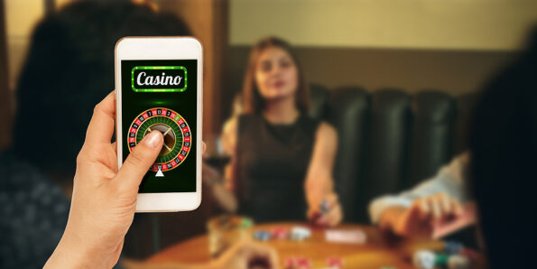 Online gambling, casino concept. Hand holding device with lottery, casino cover. Playing dips and cards on table on the background. Poker, bookmaking, gaming, modern technologies, business and finance.