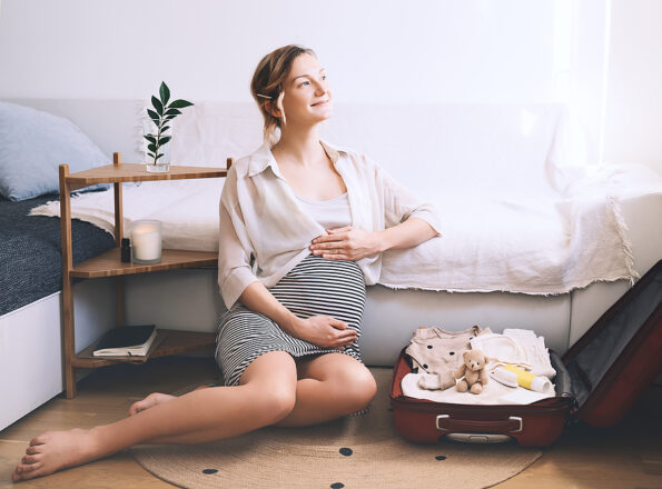 Pregnant woman is packing suitcase for maternity hospital getting ready for childbirth. Happy young mother with travel luggage of baby clothes at home. Preparation for newborn birth during pregnancy.