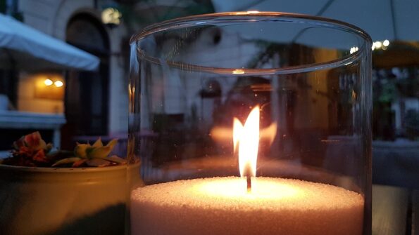 Transparent glass candle holder with flame inside. Burning fire of candle on table. Blurred reflection of glowing candle flame in glass surface. Cozy evening still life with candlestick on foreground