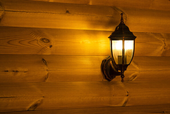 The wall mounted electric street light on the wall of a wooden log house shines yellow in the evening.Warm cozy home background blank for a postcard with a blank space for text.