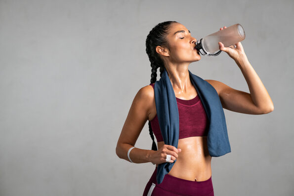 Fitness woman drinking water standing on gray background with copy space. Portrait of sweaty latin woman take a break after intense workout. Mid adult lady drink from water bottle after gym workout.
