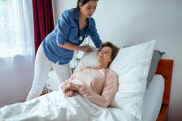 Home Nurse Making Elderly Patient Bed. Female Caregiver Adjusting Pillow For Senior Woman Lying in Hospital Bed.