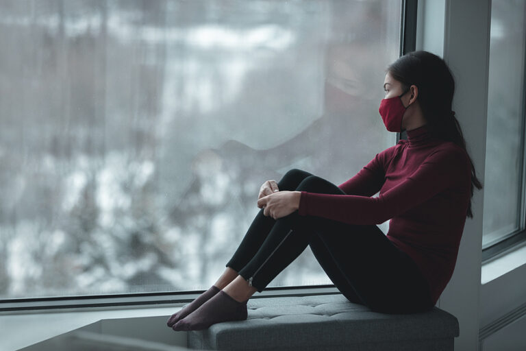 Winter depression because of coronavirus confinement. Sad Asian woman alone during city lockdown wearing face mask indoors at home covid. Anxiety, stress, mental health issues.