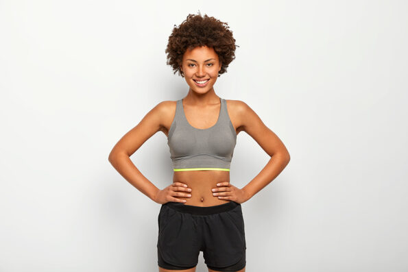 Photo of optimistic dark skinned sporty woman keeps hands on waist, smiles happily, dressed in sport bra and black shorts, isolated over white background, has fitness training with instructor