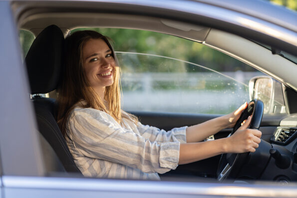 Happy young girl sitting on driver seat in new car joyful smiling hold hands on wheel. Cheerful female driving vehicle looking through open window. Successful woman car owner or getting driver license