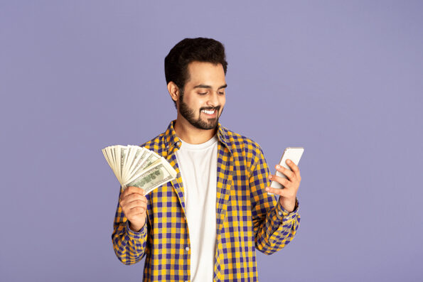 Millennial Indian guy with cash making money online on mobile phone, lilac background