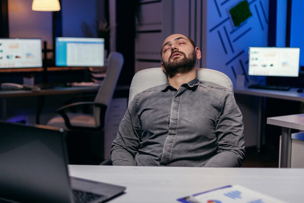 Overworked exhausted man sleeps on chair in empty office. Workaholic employee falling asleep because of while working late at night alone in the office for important company project.