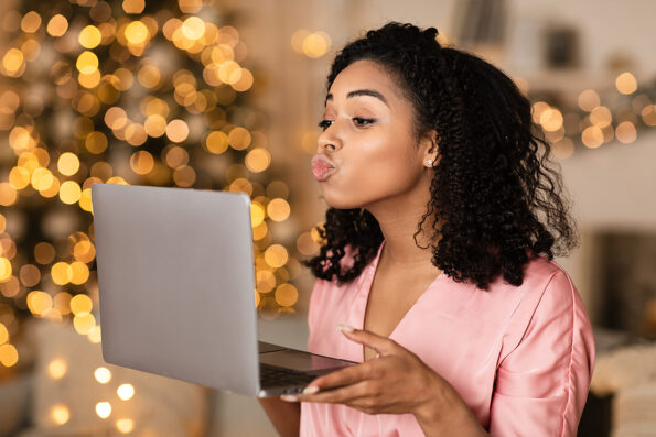 Virtual Or Long Distance Relationship And Online Dating. Portrait of beautiful young curly black woman holding laptop computer in hands and sending kisses to web camera during video chat with partner