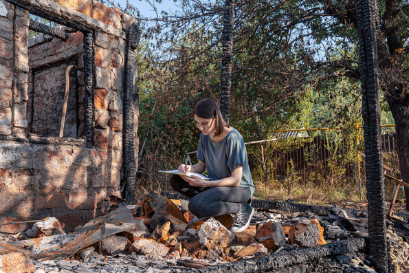 Young woman writes on the papers sitting in front of house destroyed with fire. Damaged window, walls and floor, burnt wooden frame, ash and ruins, holes in roof. Insurance agent or emergency concept. fire insurance.