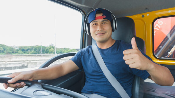 An Asian male truck driver is driving down the road with caution. He uses headphones to talk on the phone and listen to music.