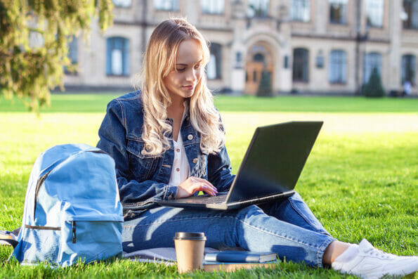 Attractive smiling student dressed casually studying outdoors on campus lawn at the university. Young woman is reading book, sitting with laptop on green grass in college park. Education concept.