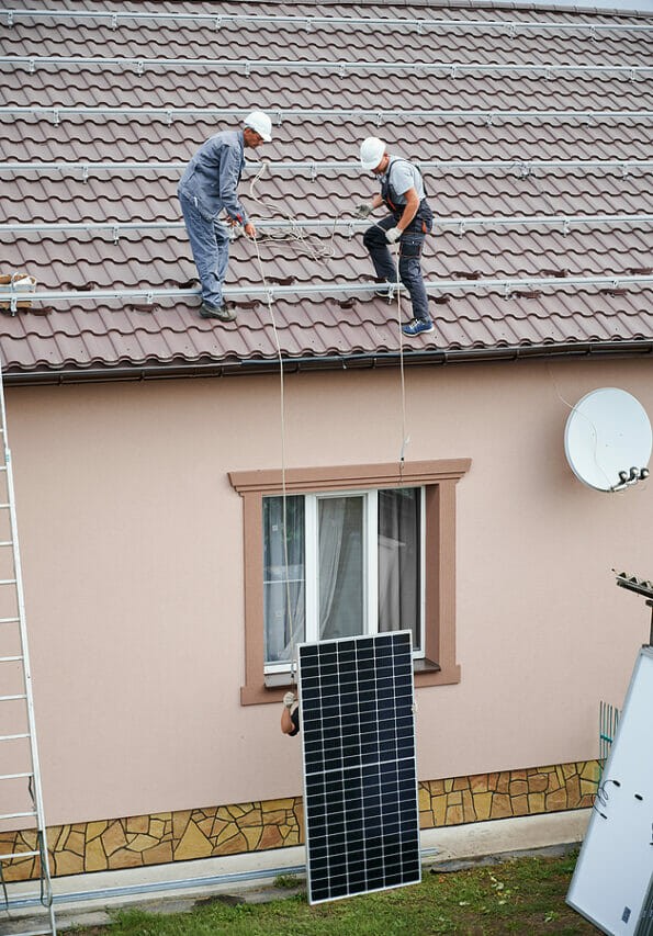 Men installers lifting up photovoltaic solar modul on roof of house. Electricians in helmets installing solar panel system outdoors. Concept of alternative and renewable energy.