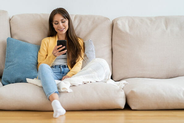 Smiling millennial woman relax sit on couch at home, using phone, texting chatting with friend. Happy girl resting on sofa hold smartphone, shopping online, check mobile application or social media.