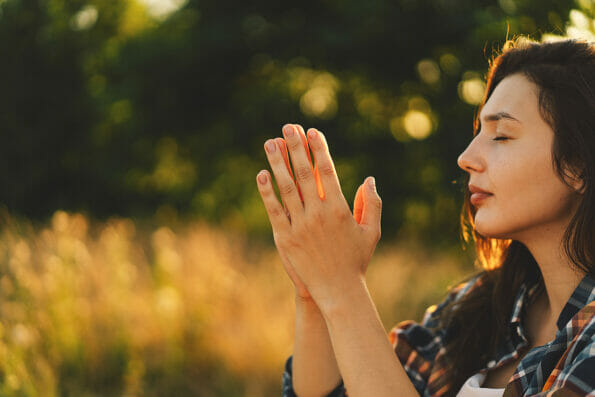 Young woman closed her eyes, praying in a field during beautiful sunset. Hands folded in prayer concept for faith, spirituality and religion. Peace, hope, dreams concept