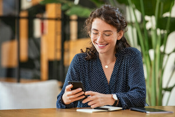 Young smiling business woman sending text message with smartphone at desk. Woman using mobile phone to check email at work with copy space. Cheerful student girl messaging online with cellphone.