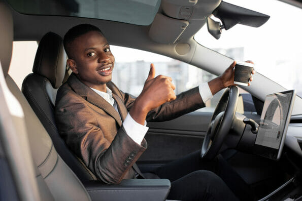 Cheerful smiling African man businessman, sitting in his new high tech electric car with upgraded avtopilot self-driving system, holding take away coffee and showing thumb up