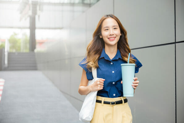 Asian beautiful business woman confident smiling with cloth bag holding steel thermos tumbler mug water glass she walking outdoors on street near modern building office, Happy female