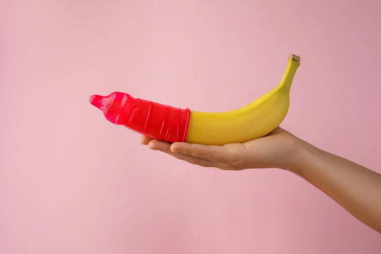 Woman holding banana in condom on pink background, closeup. Safe sex concept